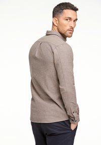 Camicia marrone a maniche lunghe con colletto, realizzata in un tessuto testurizzato, con bottoni decorativi sui polsini e un orlo dritto. Indossata con pantaloni blu navy.