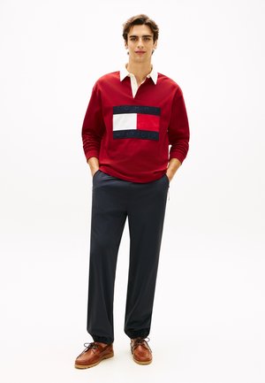 Red sweatshirt with cream collar and embroidered logo; paired with dark trousers and brown shoes. Simple design with a relaxed fit.