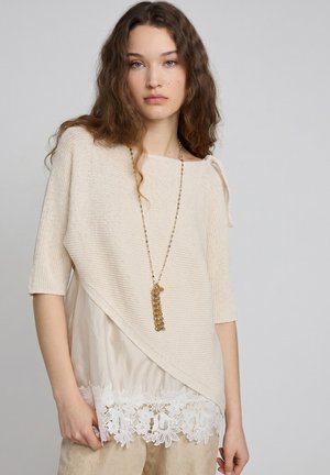 Frau mit langen lockigen Haaren, die ein beiges asymmetrisches Stricktop über einer blütenbesetzten Bluse trägt, accessoriert mit einer langen goldenen Kette.