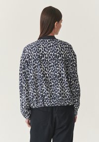 Vrouw met lang bruin haar in een losse, langemouwen blouse met blauwe en witte stippen en zwarte broek, staande tegen een effen lichte achtergrond.