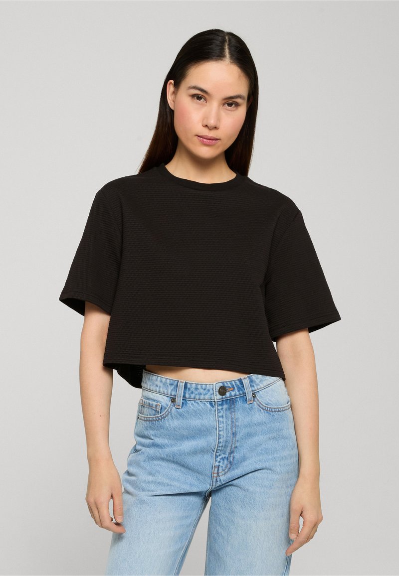 Jeune femme aux longs cheveux bruns portant un haut noir texturé à manches courtes et coupé court, et un jean taille haute bleu clair, debout devant un fond uni.