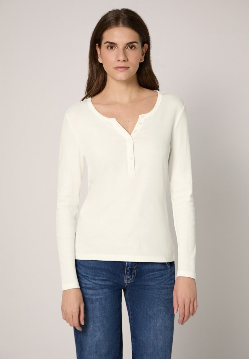 Witte top met lange mouwen, een ronde halslijn en een gedeeltelijke knoopsluiting. Zacht materiaal en een aansluitend ontwerp, gecombineerd met donkerblauwe jeans.