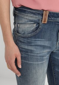 Jeans de denim azul deslavado con corte ajustado, que presentan un acento de cuero marrón claro en la cintura y un diseño de bolsillo frontal.