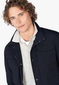 Chaqueta navy con botones a presión, cierre de cremallera y dos bolsillos en el pecho, llevada sobre un suéter gris claro. El modelo tiene el cabello rizado y una expresión neutral.