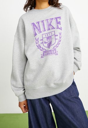 Osoba v světle šedé mikině Nike s purpurovým logem a textem "1972 Oregon" v kombinaci s tmavě modrými širokými džíny.