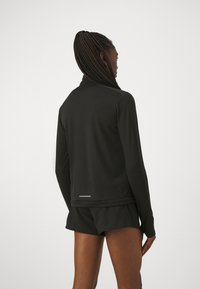 Svart träningsjacka med långa ärmar, hög krage och en reflexrand på baksidan. Tyget verkar slätt och åtsittande. Shorts är svarta.