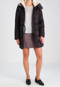 Manteau en duvet noir avec un col en fausse fourrure, fermeture éclair et pressions. Porté sur un pull gris et une jupe à motifs, accompagné de ballerines noires.