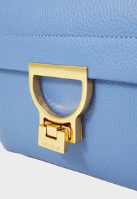 Jasnoniebieska skórzana torebka o prostokątnym kształcie, ze złotymi akcentami i charakterystycznym zapięciem z wygrawerowanym logo.