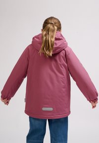 Barn med lysebrun hestehale iført en støvet lyserød hættejakke og blå jeans, vendt væk med armene let udstrakte.