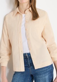 Femme portant une chemise beige clair boutonnée par-dessus un haut blanc et un jean bleu, posant devant un fond blanc, torse et jambes supérieures visibles.