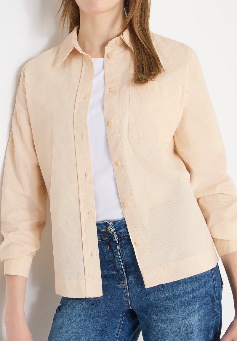 Femme portant une chemise beige clair boutonnée par-dessus un haut blanc et un jean bleu, posant devant un fond blanc, torse et jambes supérieures visibles.