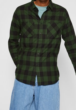 Chemise à manches longues à carreaux verts et noirs avec fermeture à boutons, deux poches poitrine et un ourlet arrondi. Portée par-dessus un t-shirt bleu clair.