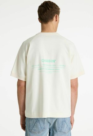 Man wearing off-white short-sleeve shirt with teal text on back, paired with light blue denim jeans, standing against a plain white background.