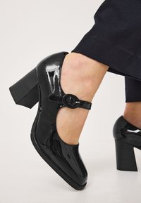 Černé lakované vysoké podpatky s kulatou špičkou, řemínkem kolem kotníku se zaoblenou sponou a robustním blokovým podpatkem.