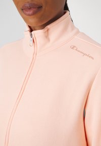 Chaqueta de color melocotón claro con cremallera, hecha de tejido suave, que presenta un cuello alto y el logo de Champion bordado en el hombro. Detalles de costura visibles.