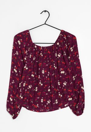 Blouse violette à manches longues avec petit imprimé floral rouge et blanc, encolure froncée et poignets élastiques, suspendue sur un cintre noir.