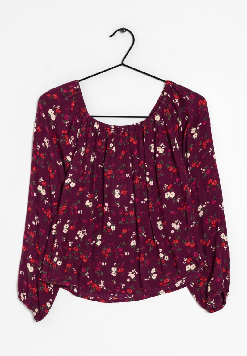 Blouse violette à manches longues avec petit imprimé floral rouge et blanc, encolure froncée et poignets élastiques, suspendue sur un cintre noir.