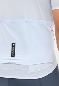 Λευκό αθλητικό πουκάμισο από δίχτυ και μονόχρωμο ύφασμα με μια μικρή μαύρη ορθογώνια ετικέτα στο κάτω μέρος της πλάτης, φορεμένο με παντελόνι σκούρου γκρι χρώματος.