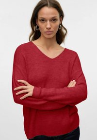 Pull en tricot rouge à côtes avec un col en V et des manches longues. Le tissu a une texture douce et une coupe décontractée. Assorti à un jean foncé.