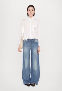 Witte blouse met lange mouwen en een voorzaklogo, gecombineerd met lichtblauwe wijde jeans. Draagt een kleine crèmekleurige schoudertas.