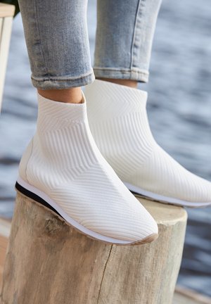 White ankle boots with a textured ribbed pattern, soft material, and a flexible design. Features a slim sole with a contrasting black edge.