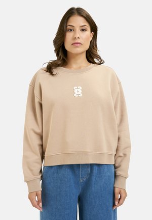 Beige cropped sweater with a round neckline and long sleeves, featuring a textured white logo at the center, paired with blue denim.