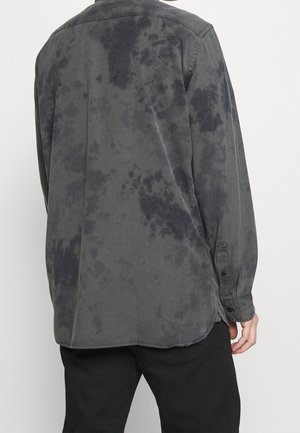 Vue de dos d'une personne portant une chemise à manches longues gris foncé avec des motifs noirs irréguliers et un pantalon noir sur un fond clair uni.