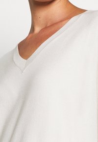 Pull blanc à col en V en matière tricotée douce, avec une coupe décontractée et des accents côtelés subtils le long de l'encolure et de l'ourlet.