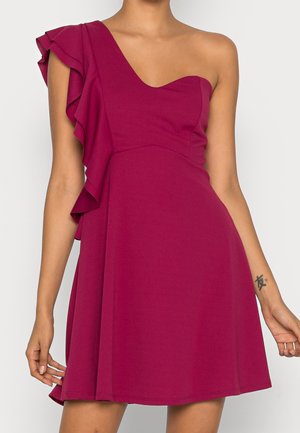 Woman wearing a sleeveless magenta dress with a ruffle detail on one shoulder and an asymmetrical neckline.