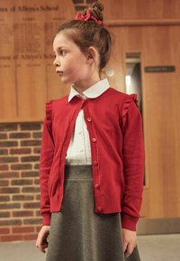 Red cardigan with ruffled shoulder details, buttoned front; paired with a white collared shirt and gray skirt. Background with wooden panels.