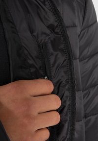 Veste matelassée noire avec une texture lisse et brillante, une poche zippée sur le côté, une silhouette ajustée et des détails de coutures.