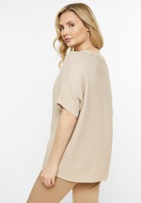 Beige pletený svetr s krátkým rukávem, volným střihem, ribbovanou texturou a výstřihem do kulata. Zadní část má lehce prodloužený lem.