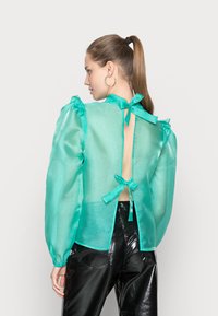 Blusa translúcida turquoise com mangas bufantes, laços na parte de trás do pescoço e detalhe nas costas descobertas; combinada com calças pretas brilhantes.