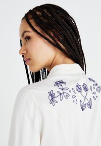 Hvit skjorte med en blomsterbroderi design i mørkeblått på ryggen. Skjorten har en klassisk krave og en glatt tekstur.