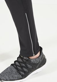 Svarta träningsbyxor med en sidoficka med dragkedja och reflexdetaljer, tillsammans med grå och svarta mönstrade sneakers med svarta accenter.