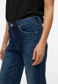 Kõrge vöökohaga sinised teksapüksid, helepesuga, esitaskute ja kontrastsete õmblusedetailidega. Sile tekstuur ja keha järgiv lõige.