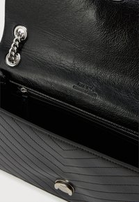 Mustmust mustkott, millel on tikandiga mustriga nahk, hõbedased detailid ja tõmblukuga tasku. Tootel on ka sisetaskus logo sisse pressitud.