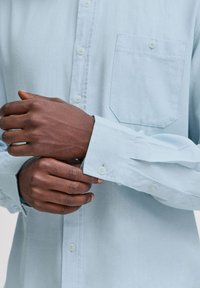 Cămașă button-up, cu mâneci lungi, de culoare albastru deschis, având un buzunar pe piept, butoane albe și detalii de coasere vizibile pe manșete.