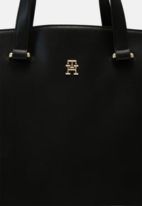 Sac à main en cuir noir avec double poignée et un petit emblème logo en métal doré centré près du haut.