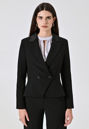 Schwarzer maßgeschneiderter Blazer mit doppelt geknöpften Vorderseite, Reverskragen und strukturierter Silhouette, kombiniert mit einem hellgrauen Oberteil mit hohem Kragen.