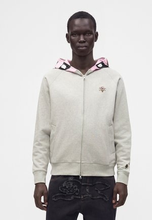 Mann trägt hellgrauen Kapuzenpullover mit Reißverschluss und rosa-schwarz gemustertem Kapuzenfutter sowie dunkle Hose mit strukturierten Designs vorne.
