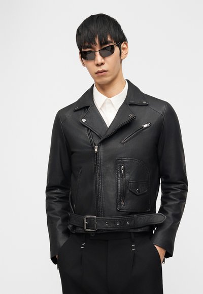 Άνδρας που φοράει μαύρο δερμάτινο μπουφάν μοτοσικλετιστή, λευκό πουκάμισο με κουμπιά, μαύρο παντελόνι και σκούρα γυαλιά ηλίου, στέκεται με τα χέρια στις τσέπες.