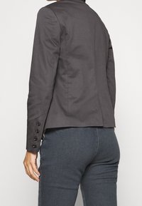 Szary dopasowany blezer o gładkiej fakturze, z klapą z wycięciem oraz pięcioma guzikami na rękawach; krój z pionowymi przeszyciami.