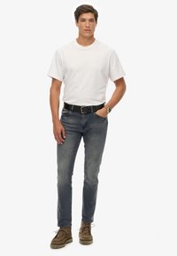Camiseta blanca de algodón, jeans azul desgastados, cinturón negro y zapatos de ante marrón. El modelo está de pie, con las manos en los bolsillos, mirando hacia la cámara.