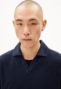 Marineblauwe pullover van gestructureerde stof, met een V-vormige kraag en korte mouwen, gedragen over een zwart kledingstuk. Gladde oppervlakte, aansluitend ontwerp.