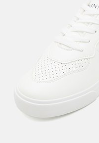 Hvit sneakers med en glatt syntetisk overdel med perforerte detaljer og flate lisser; gummisåle med teksturert grep.
