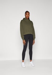 Olivgrüner, verkürzter Hoodie mit Logo auf der Vorderseite, kombiniert mit schwarzen Leggings und weißen Sneakers. Glatter Stoff, lässige Passform und Kapuzen-Design.