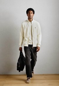 Hvid zip-up jakke, sorte jeans og beige sneakers. Bærer en sort taske. Glat stof og enkelt design, sat mod en lys baggrund.