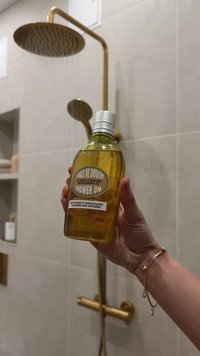 Bottiglia di olio per doccia in vetro trasparente con liquido giallo, tappo argentato ed etichetta con scritta nera "Huile de Douche", tenuta davanti a una doccia.