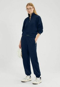 Felpa con zip di colore blu navy e colletto alto, abbinata a pantaloni jogging coordinati. Il materiale sembra morbido; presenta un elastico in vita e tasche laterali.
